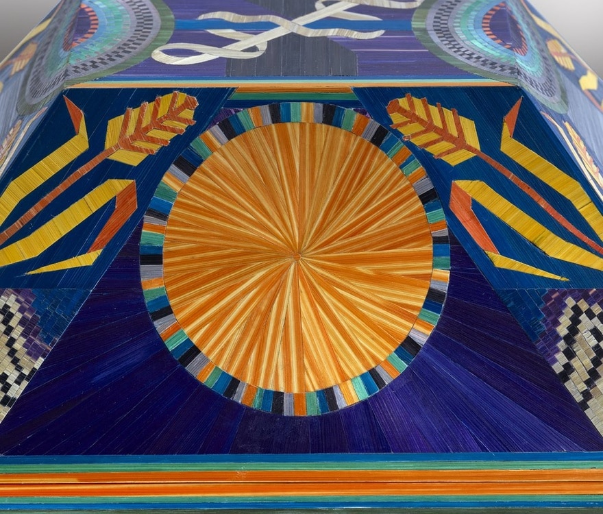 Coffret orné de motifs décoratifs et narratifs : le soleil, symbole de lumière et de prospérité, le blé, référence directe à l'artisanat, et quatre pieds cylindriques évocateurs d'un tabernacle.