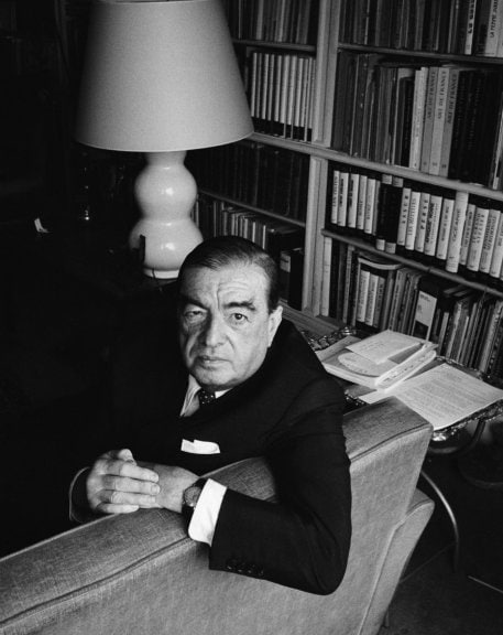 Roger Caillois is sitting in a couch. Right behind him there is a library. Picture is in black and white